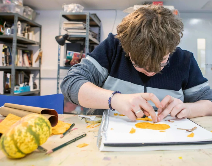 Student working on an art project, using orange paper to create a design in a sketchbook, surrounded by art supplies. Student working on an art project, using orange paper to create a design in a sketchbook, surrounded by art supplies.